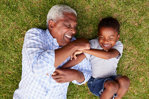 A grandfather and grandson lie on a lawn, smiling as they embrace each other.
